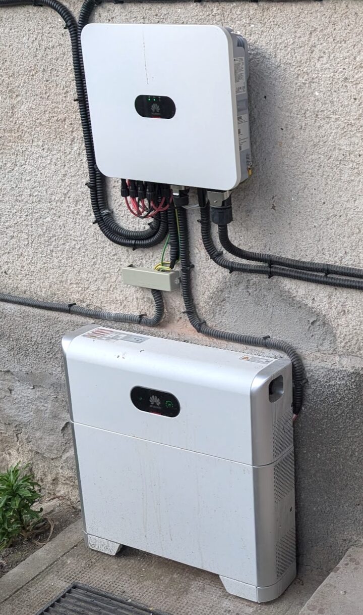 Un invertor Huawei SUN2000 alb pe un perete exterior, cu o baterie LUNA2000 așezată pe jos sub el